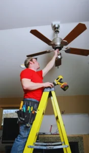 Electrician installing a ceiling fan while standing on a ladder with tools and drill